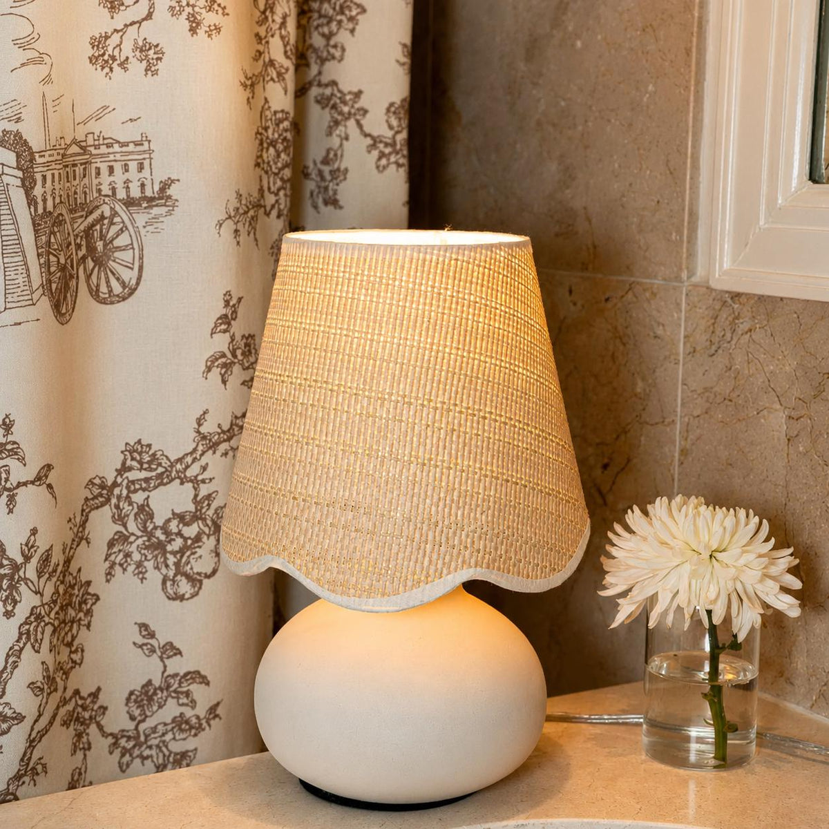 Small woven shade lamp with a rounded ceramic base styled beside flowers in a warm interior.