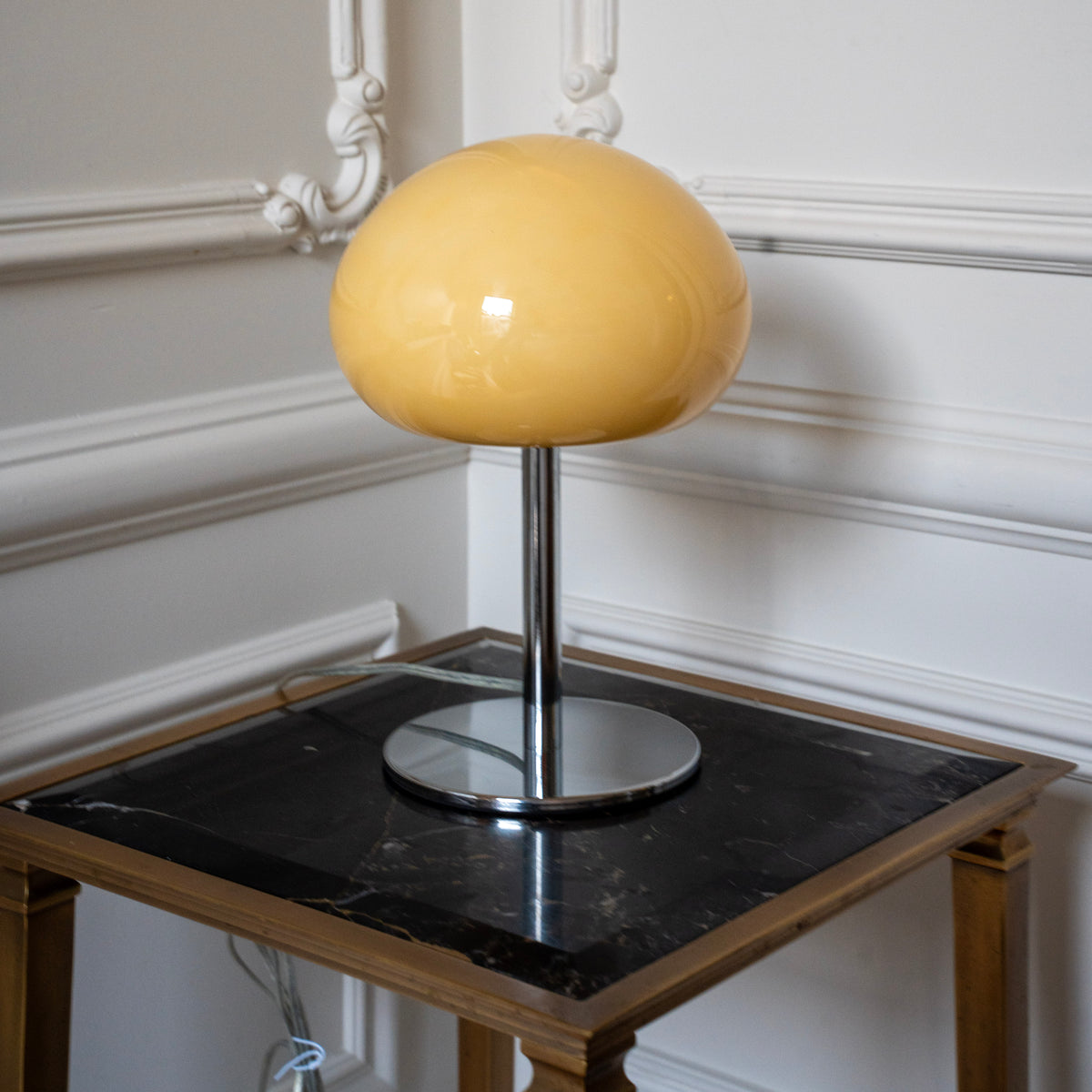 Sculptural cream glass lamp styled on a black marble side table against decorative white wall moulding.