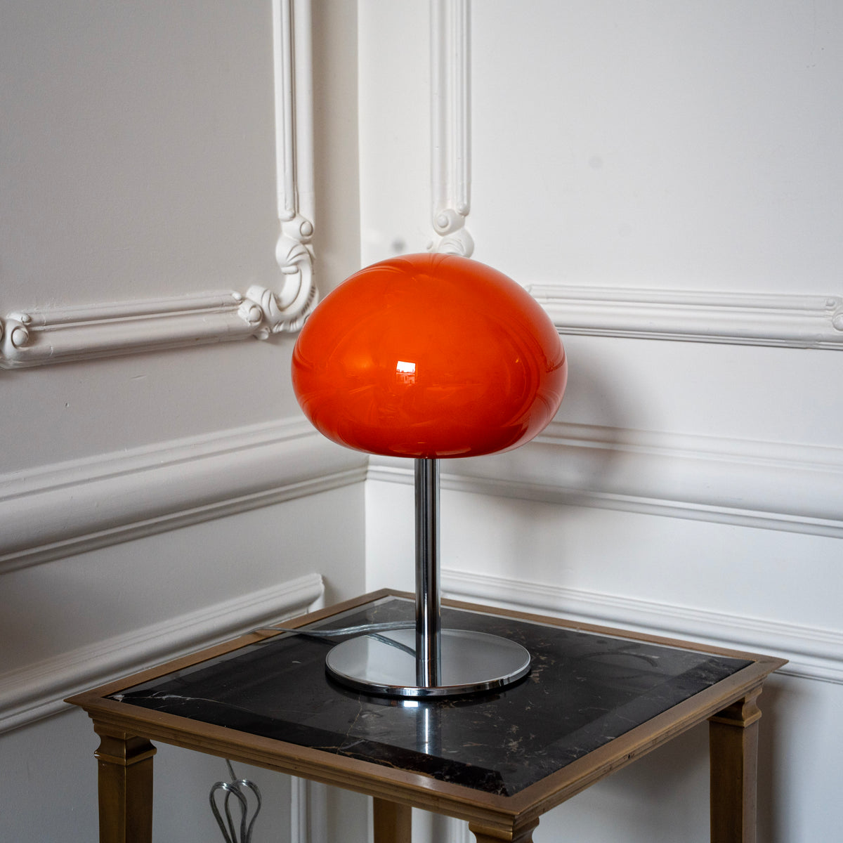 Orange mushroom table lamp with chrome base styled against ornate white wall moulding in a classic interior.