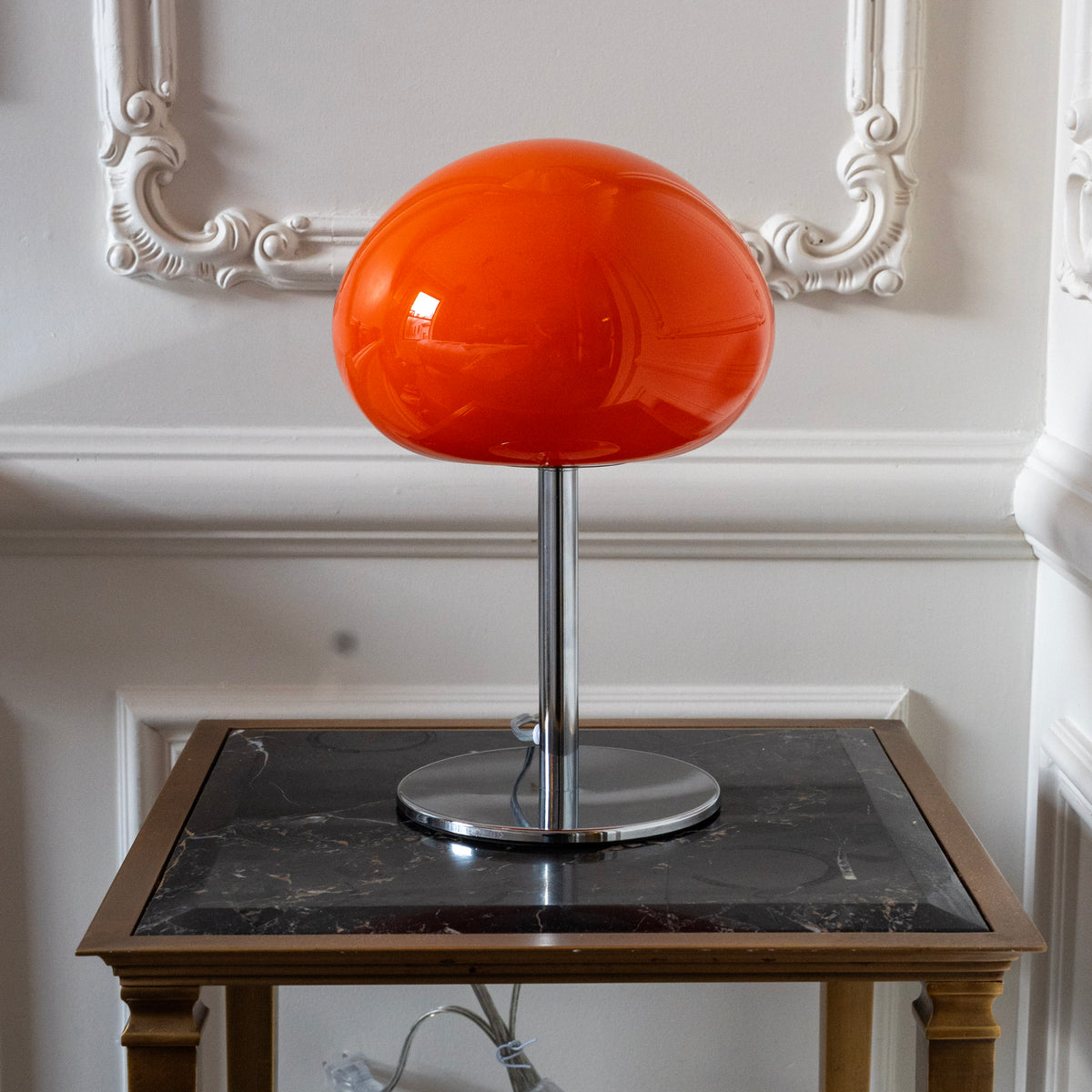 Sculptural orange glass table lamp styled on a black marble side table against decorative white wall moulding.
