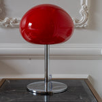 Red mushroom table lamp with chrome base styled against ornate wall moulding in a classic neutral interior.
