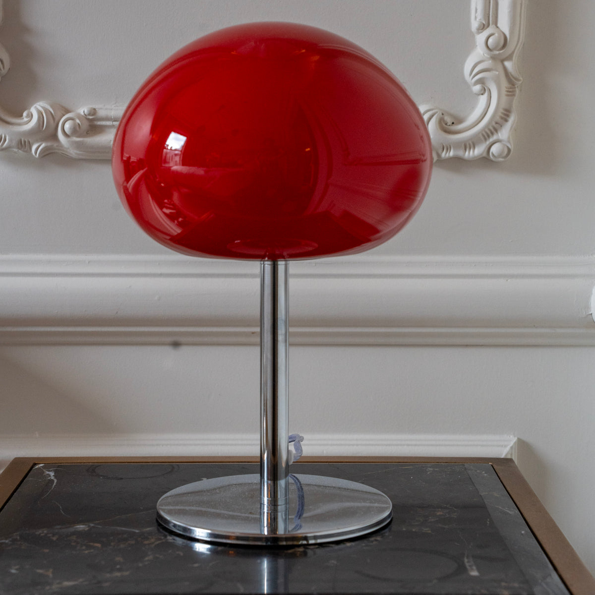 Red mushroom table lamp with chrome base styled against ornate wall moulding in a classic neutral interior.
