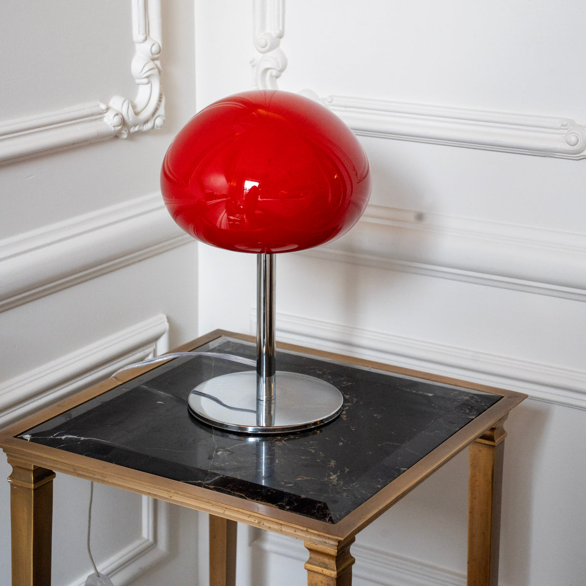Sculptural red glass table lamp styled on a black marble side table against decorative white wall moulding.