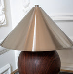 Close-up of a brushed brass cone shade above a rounded woodgrain lamp base against white wall moulding.
