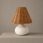 Small ceramic table lamp with a rounded cream base and woven rattan shade on a neutral studio backdrop.