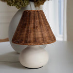 Rattan and ceramic table lamp styled beside a window in a softly lit neutral interior.