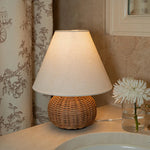Warm rattan table lamp styled in a softly lit bedroom with a white fabric shade.