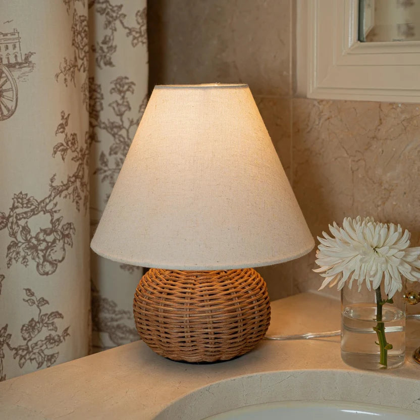 Warm rattan table lamp styled in a softly lit bedroom with a white fabric shade.