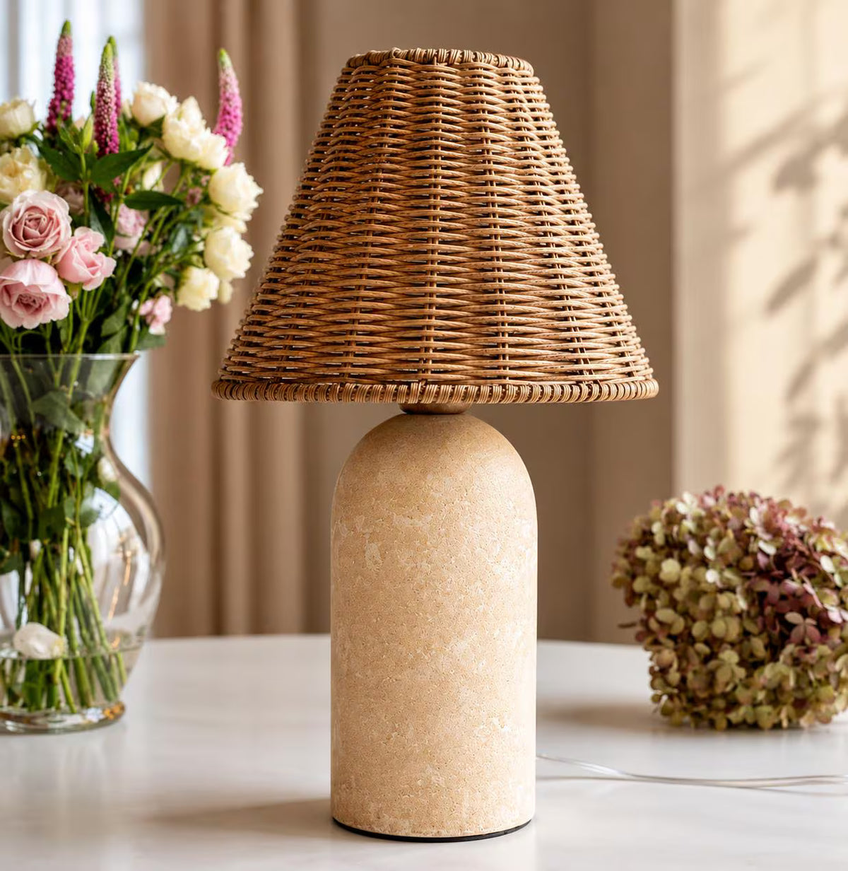 Rattan and sandstone table lamp styled beside flowers in a warm, collected interior.
