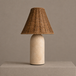 Small rattan table lamp with a sandstone ceramic base and woven shade on a neutral pedestal.