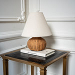 The Reed table lamp with a woven rattan base and off-white fabric shade styled on a marble and brass side table.