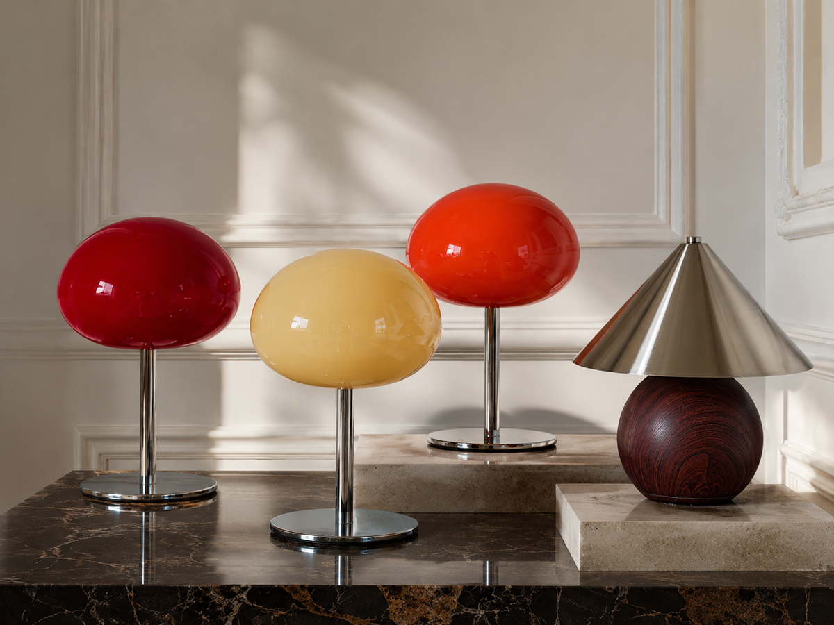 Four decorative table lamps displayed on a dark marble surface against an elegant cream paneled wall. Three lamps have glossy spherical shades in red, pale yellow, and orange on chrome stands, while a fourth lamp features a conical brushed-metal shade resting on a dark wood sphere. Sculptural table lamps in a warm interior